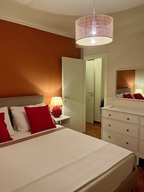 Habitación de hotel con cama, mesita de noche, lámpara, cómoda y una puerta que da a otra habitación.