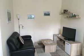 Een woonkamer met een zwarte leren bank, een salontafel, een televisie op een houten standaard, en een wandplank met boeken.