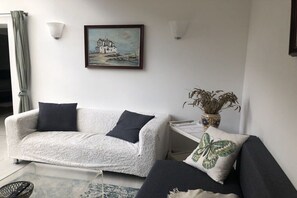 Ein Wohnzimmer mit einem weißen Sofa, einem schwarzen Sofa, einem Gemälde einer Küstenlandschaft, einer Vase mit Trockenpflanzen und einem kleinen Tisch.