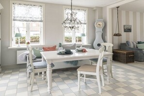 Ein Essbereich mit einem weißen Tisch, Stühlen und einer Kristallleuchter. An der Wand hängt eine Uhr, und ein Fenster ist mit Vorhängen versehen.