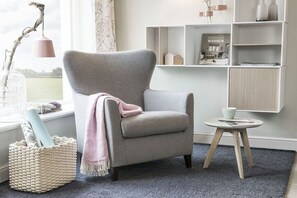 Ein Zimmer mit grauem Sessel, einem kleinen runden Tisch, einem gewebten Korb und einem Bücherregal mit Dekoration.