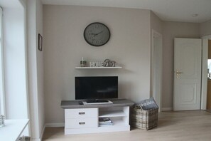 Ein Zimmer mit Fernseher auf einem weißen Schrank, einer Uhr an der Wand und einer Tür zur rechten Seite.