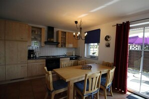 A kitchen with wooden cabinets, a dining table with chairs, a chandelier, and a window with curtains. - Holiday home with 1 bedroom (Poel \/ OT Kaltenhof)