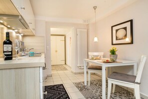A modern kitchen with a white countertop, a wine bottle, and a glass. There is a dining area with a white table and chair, a potted plant, and a framed picture on the wall. - Holiday apartment in Binz with swimming pool (Binz)