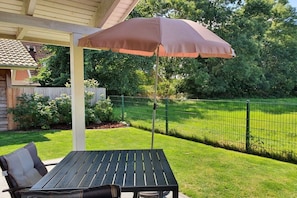 Eine Terrasse im Freien mit Tisch und Stühlen, einem braunen Sonnenschirm und einem grünen Zaun.