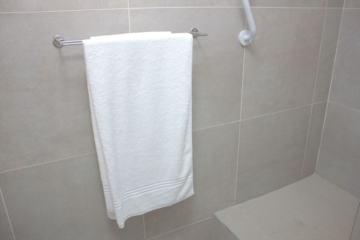 Una toalla blanca colgada en un perchero de metal en un baño con paredes de azulejos y una zona de ducha.