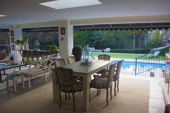 A dining area with a table and chairs, a glass coffee table, and a view of a swimming pool and garden.