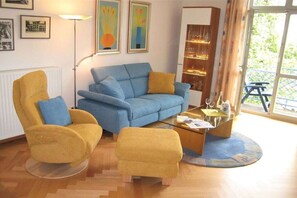 Una sala de estar con un sofá azul, un sillón amarillo y un reposapiés, una mesa de centro de vidrio y una estantería.