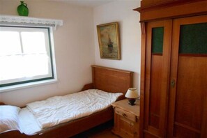 Ein Einzelbett mit weißer Matratze und Holz-Kopfteil, ein Nachttisch mit Lampe, ein grüner Blumenvasen auf der Fensterbank und ein gerahmtes Bild an der Wand.