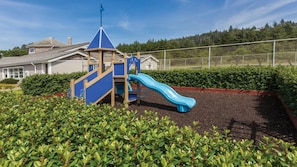 Un parque infantil con un tobogán azul y una estructura para escalar, rodeado por una valla y follaje.
