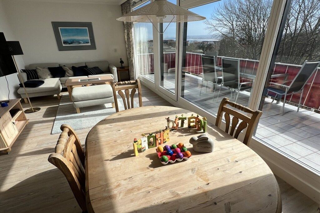 A dining area with a wooden table and chairs, a view of the outdoors through large windows, and a sofa in the background.