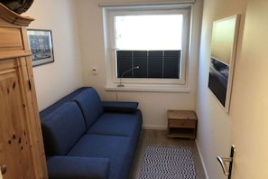 Ein kleines Zimmer mit einem blauen Sofa, einem Fenster mit Jalousien, einem kleinen Holztisch und einem gemusterten Teppich.