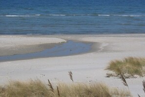 Une plage de sable avec de l’eau, et quelques touffes d’herbe au premier plan.
