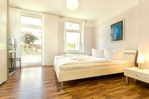 A bedroom with a bed, a nightstand, a lamp, a TV, and a balcony view. - Holiday apartment in Binz with swimming pool (Binz)