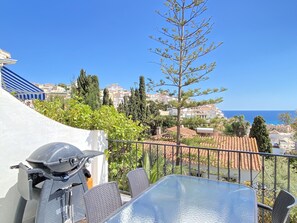 A balcony with a grill, a table, and chairs, overlooking a coastal town with buildings and a clear sky. - Villa in Nerja Near Burriana Beach (Nerja)