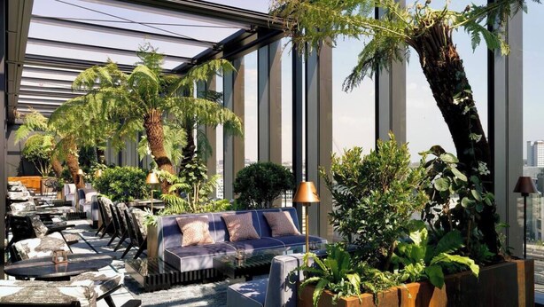 Un moderno salón en la azotea con techo de vidrio, asientos al aire libre y abundante vegetación.