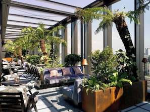 A modern rooftop lounge with a glass ceiling, outdoor seating, and lush greenery.
