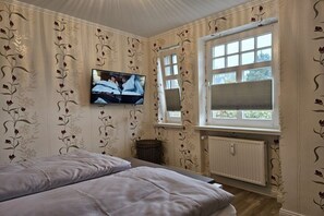 Ein Schlafzimmer mit einem Bett, einem an der Wand montierten Fernseher, einem Fenster mit Jalousien und einer Heizung.
