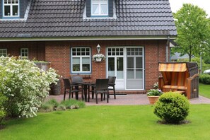 A brick house with a tiled roof, a front porch with a table and chairs, and a small garden with a bush and flowers. - Holiday apartment with 1 bedroom (Wyk auf Föhr)