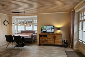 Ein Wohnzimmer mit Holzboden, einem Fernseher auf einer Holzkommode, einem Esstisch mit Stühlen und einem Sofa.