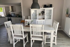 Ein Essbereich mit weißem Tisch und Stühlen, eine Vitrine mit Glastüren und eine Küche im Hintergrund.