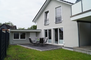 Ein zweistöckiges Haus mit einer Terrassenfläche, einem Esstisch mit Stühlen und einem kleinen Garten.