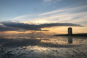 Ein Strand bei Sonnenuntergang mit ruhiger See, ein hohes Gebäude rechts und ein klarer Himmel.