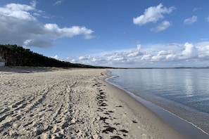 Hiekkainen ranta, jossa on tyyni meri, kirkas taivas ja taustalla metsäinen alue.