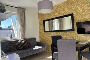 A room with a sofa, a dining table, chairs, a television, and a window with curtains. - Holiday apartment with 1 bedroom (Ostseebad Baabe)
