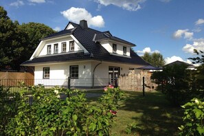 Une maison à deux étages, avec une façade blanche et un toit foncé, entourée d’une cour clôturée et agrémentée de verdure.