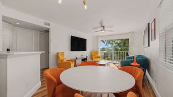 Un coin repas avec une table ronde et des chaises orange, un téléviseur fixé au mur et un ventilateur de plafond.