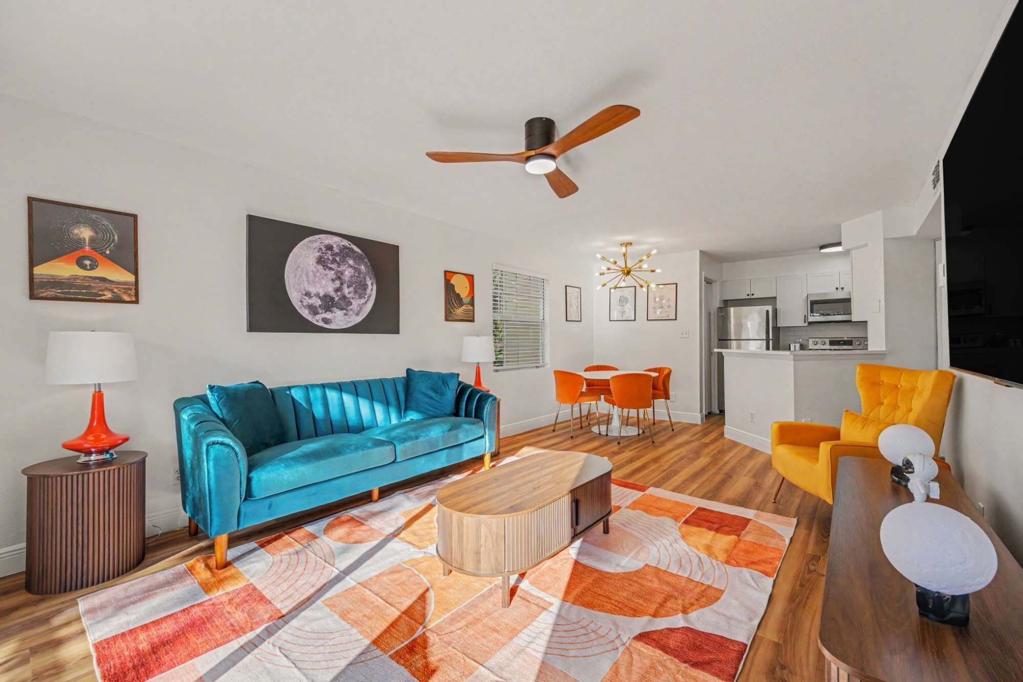 A modern living room with a blue sofa, orange chairs, a wooden coffee table, and a ceiling fan.