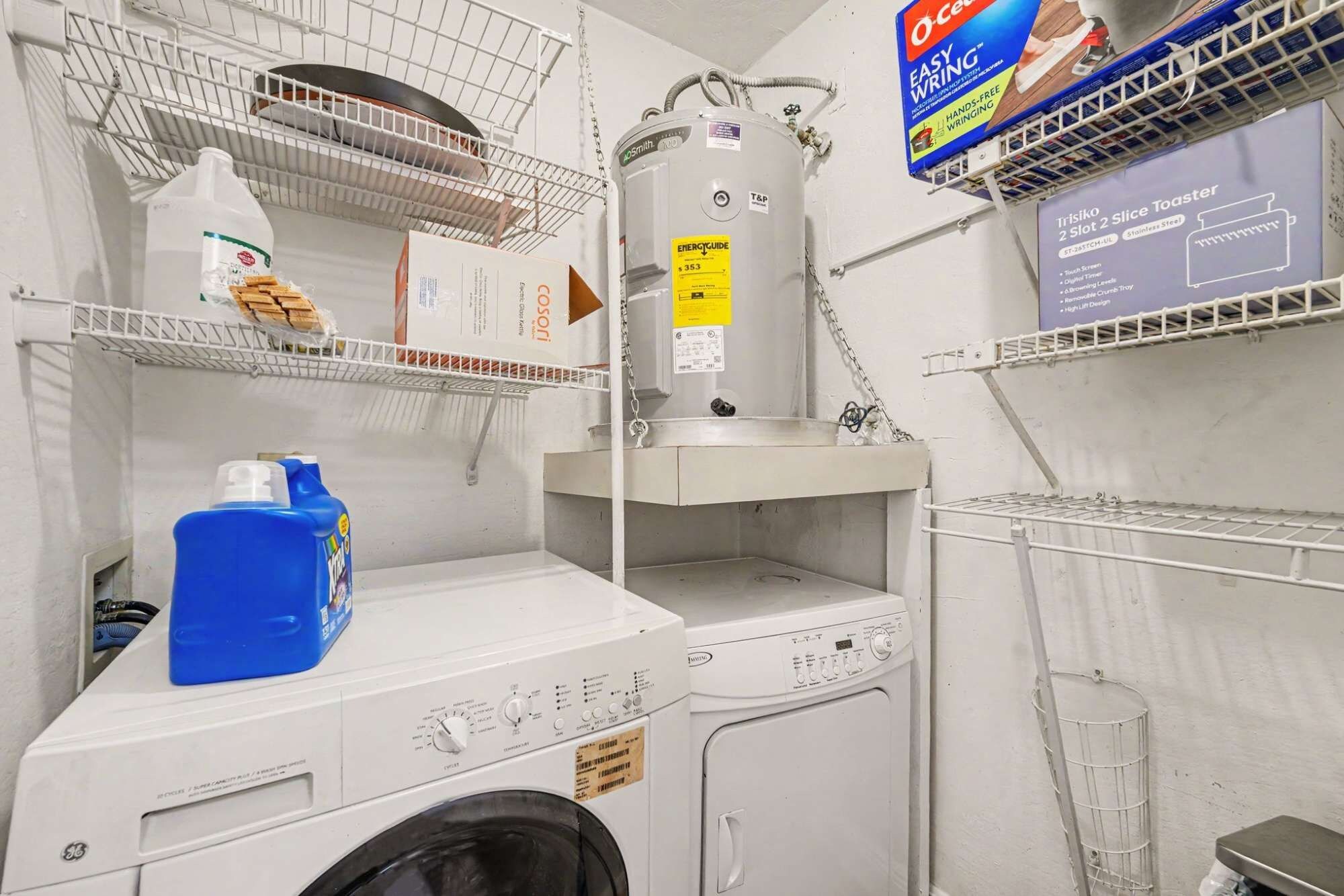 Uma lavanderia com máquina de lavar, secadora, prateleiras com diversos objetos e um aquecedor de água.