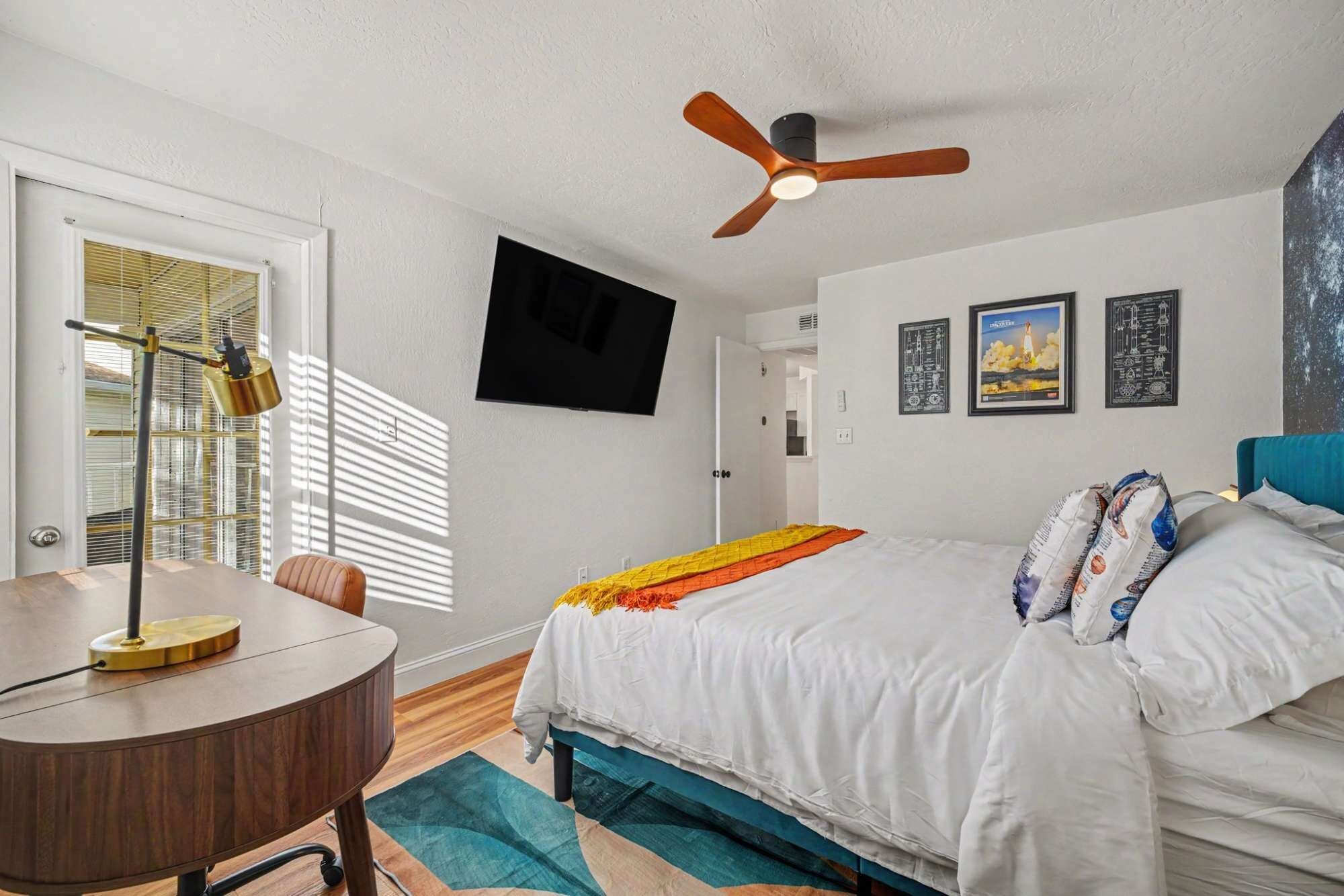 A bedroom with a bed, a desk, a ceiling fan, and a window with blinds.