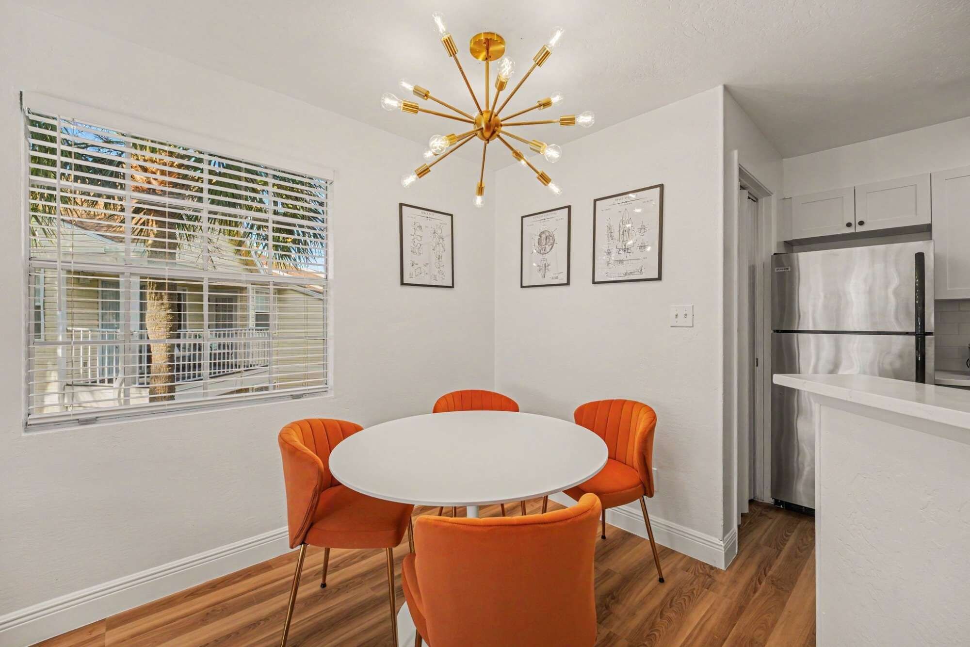 Een eethoek met een ronde tafel en vier oranje stoelen, een moderne kroonluchter, en een keuken met een roestvrijstalen koelkast.