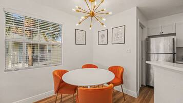 Een eethoek met een ronde tafel en vier oranje stoelen, een moderne kroonluchter, en een keuken met een roestvrijstalen koelkast.