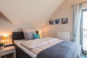 A bedroom with a bed, bedside tables, a nightstand, a window with curtains, and wall art. - Holiday apartment with 2 bedrooms (Heiligenhafen)