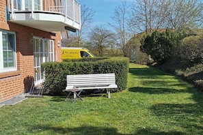 Un edificio residencial con un banco y un seto al frente, y una furgoneta amarilla al fondo.