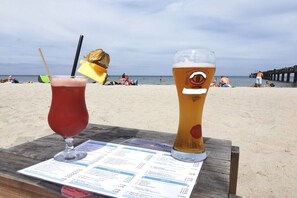 Zwei Getränke auf einem Tisch, davor eine Speisekarte vor Strand und Steg.