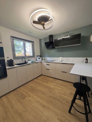 A modern kitchen with a built-in oven, a microwave, and a sink. There is a window with a view of trees outside. - Villa du Betey 4 étoiles 8pers climatisée Andernos les bains (Andernos-les-Bains)