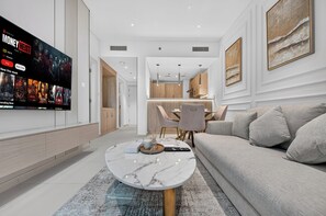 A modern living room with a flat-screen TV mounted on the wall, a marble coffee table, a sofa, and a dining area with a kitchen in the background. - Seven Palm | 1BR, Sweet, Stylish & Serene (Dubai)