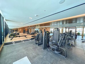 A gym with various exercise machines, a mirrored wall, and a view of the outside. - Trillionaire | 1BR, Trendy, Timeless & Tempting (Dubai)
