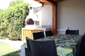 An outdoor kitchen area with a brick oven, a dining table with chairs, and a wooden fence. - Beach House in Sardinia - 100 meters from the sea, with a garden and barbecue (Quartu Sant'Elena)