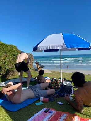 Escena de playa con gente relajándose bajo un paraguas azul y blanco, una tabla de surf y una silla de playa.