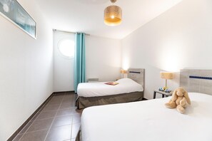 Une chambre d’hôtel avec deux lits, une fenêtre avec des rideaux, une table de chevet avec une lampe et un ours en peluche sur le lit.