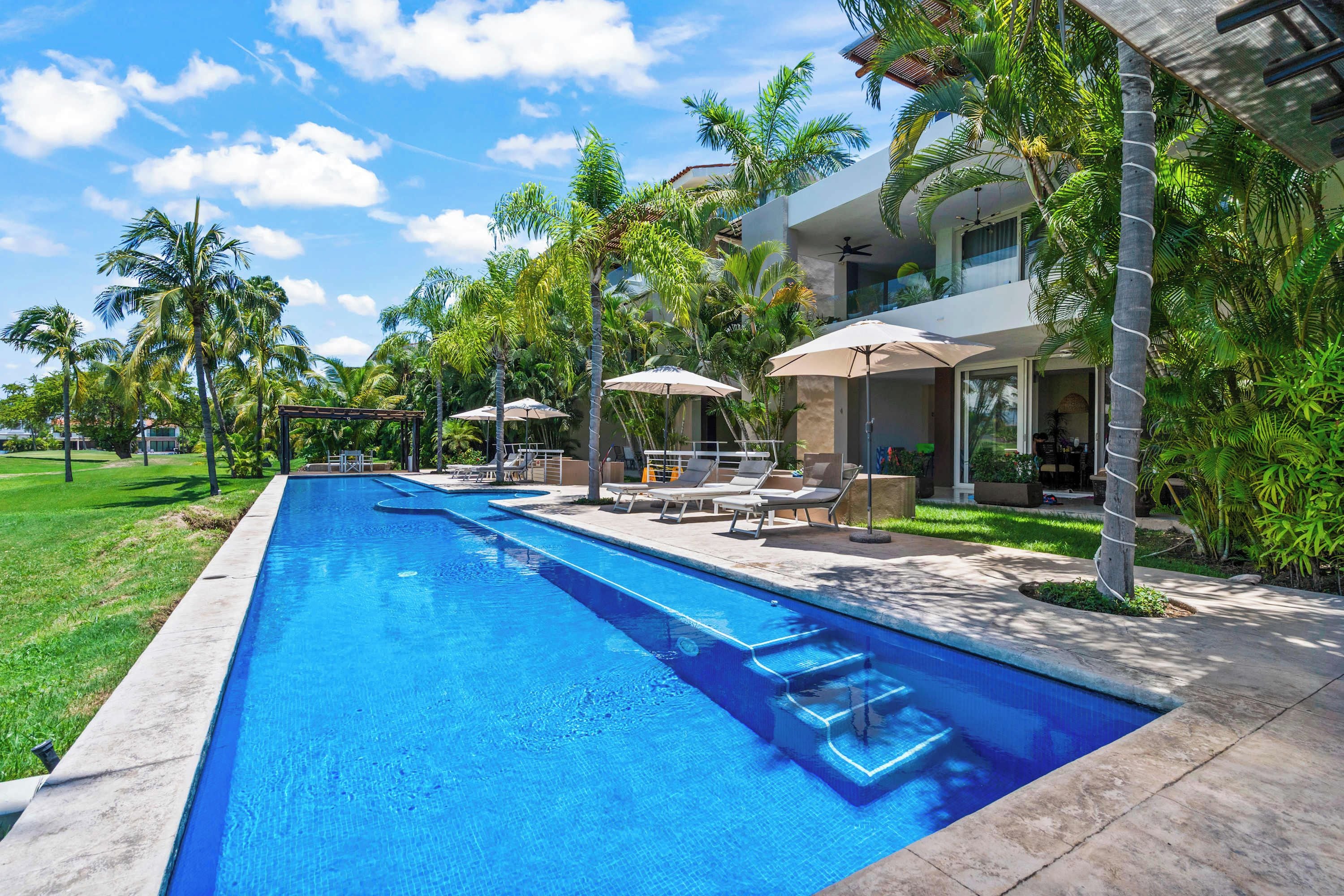 Piscina con sillones de playa y sombrillas, rodeada de palmeras y un edificio con balcón.