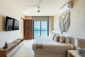 Une chambre à coucher comprenant un lit, une télévision, un ventilateur de plafond, une grande fenêtre donnant sur la mer et un climatiseur fixé au mur.