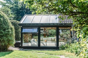 A modern glass-enclosed structure with a metal roof, surrounded by lush greenery. - Farmhouse by the sea || Langballig (Langballig)