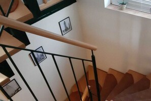 Eine Treppe mit hölzernem Geländer und schwarzen Metallspindeln, die zu einer Zwischenebene mit einem kleinen Fenster und einem Blumentopf führt.