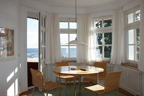 A room with a round table and chairs, large windows with curtains, and a view of the sea. - Holiday apartment with sea view, Villa Vineta 14 (Seebad Bansin)
